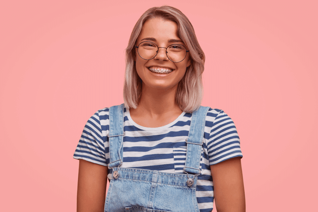 Smiling young woman with glasses showing her braces against a pink background